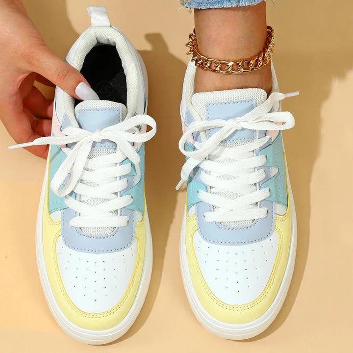Top view of pastel yellow, blue, and white casual sneakers with white laces, worn on foot with gold chain anklet