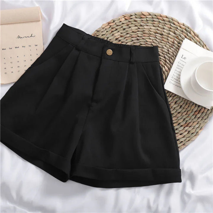 Black high-waisted pleated shorts with button detail, styled on a white surface with a woven mat, calendar, and cup