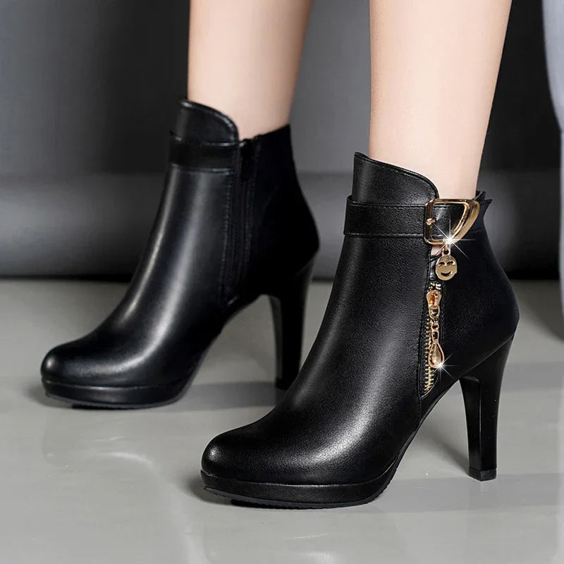Black high-heeled ankle boots with gold buckle and zipper embellishments on gray floor