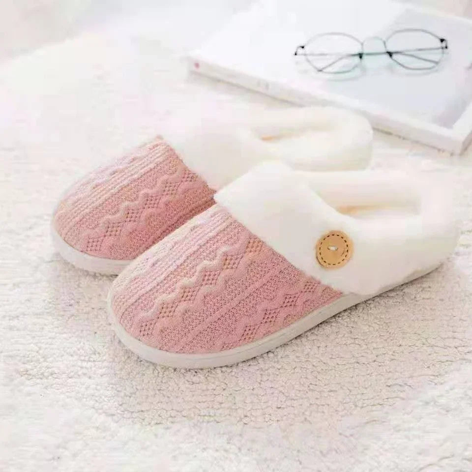 Pink knitted indoor slippers with white fleece cuffs and button detail on textured white surface