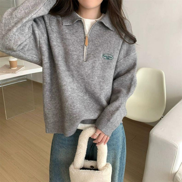 Woman wearing grey quarter-zip sweater with embroidered logo, holding white fluffy handbag in modern room