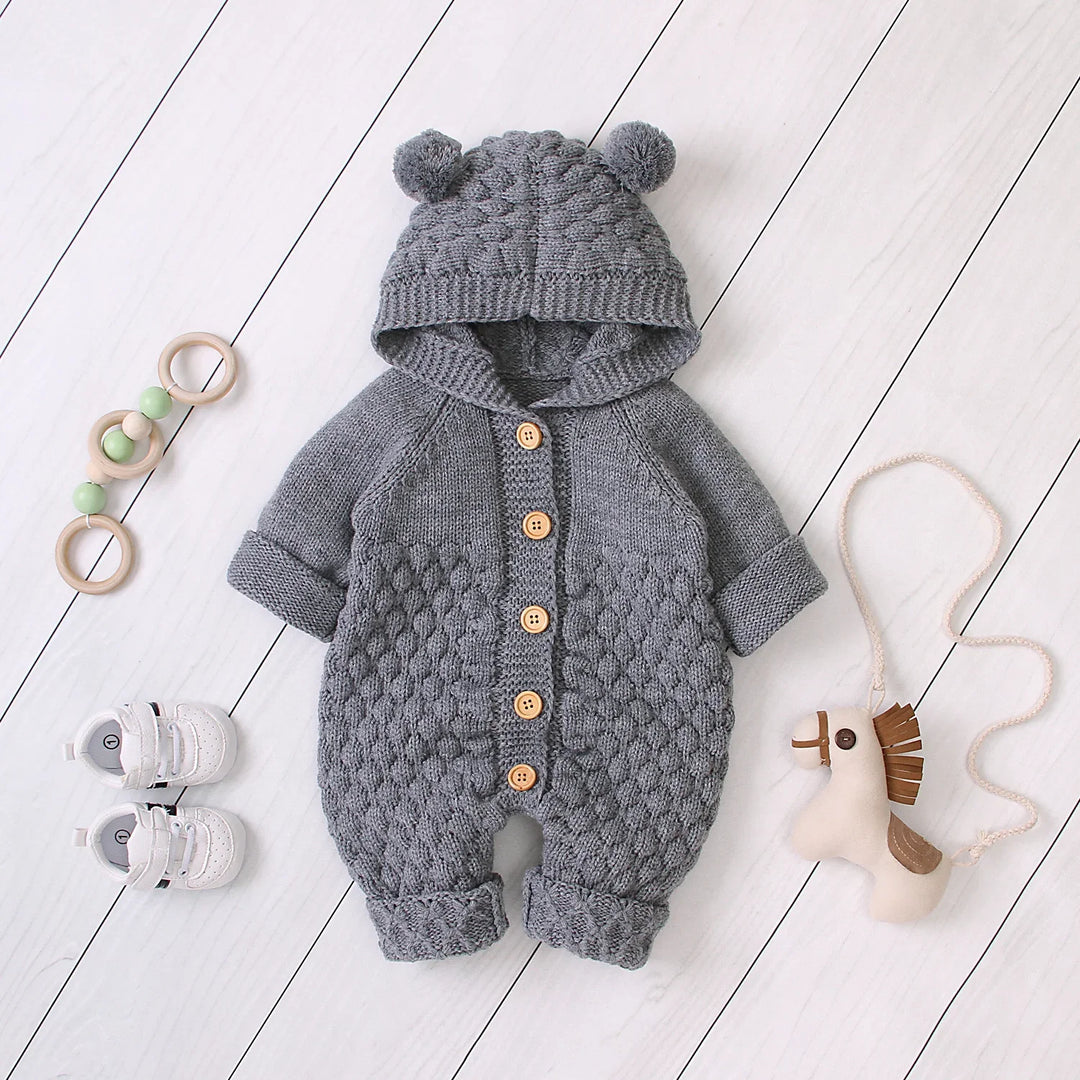 Gray knitted baby romper with hood and wooden buttons, baby shoes, toy horse, and wooden ring toy on white floor