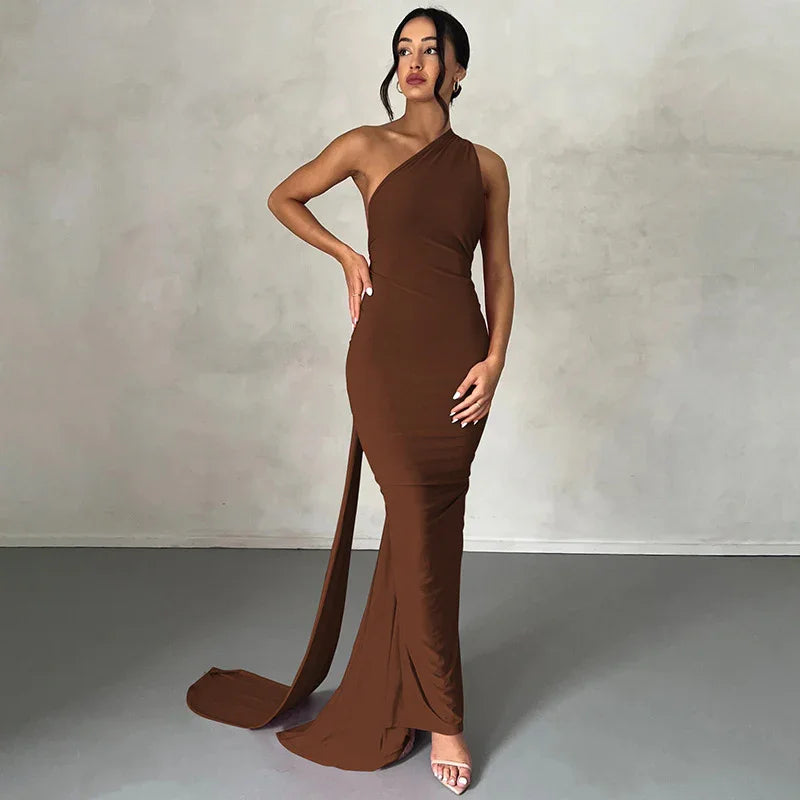 Woman wearing a brown one-shoulder fitted evening gown with a long train against a plain wall backdrop