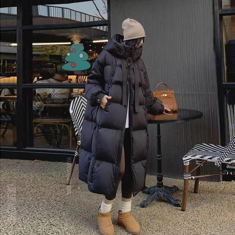 Woman in long black puffer coat, beige beanie, and tan boots standing outdoors near café table and chairs