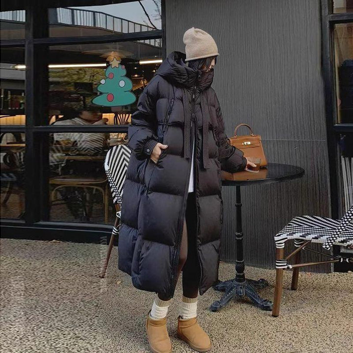 Woman in long black puffer coat, beige beanie, and tan boots standing outdoors near café table and chairs