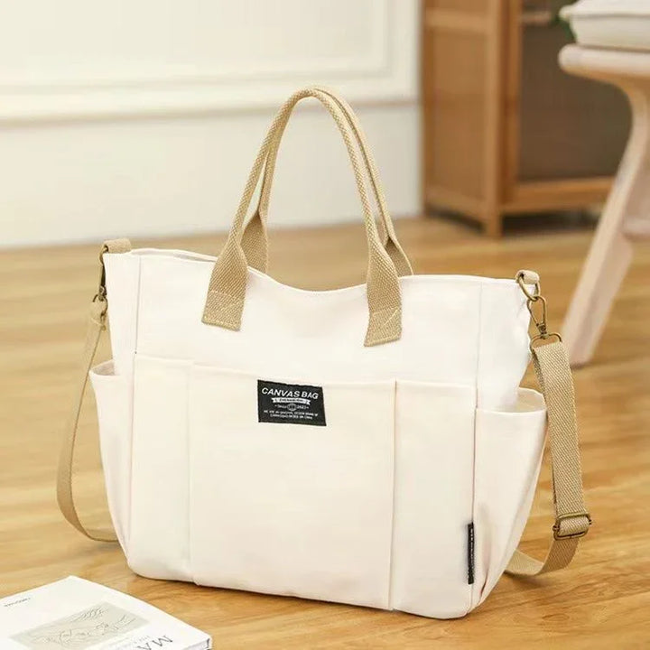 White canvas tote bag with beige handles and adjustable shoulder strap on wooden floor