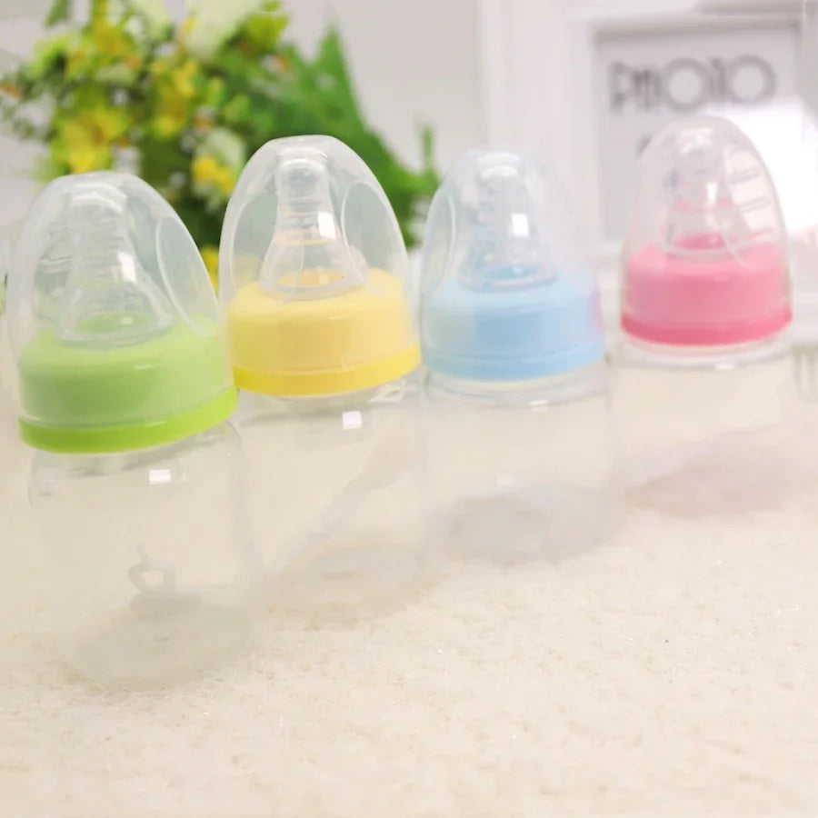 Four clear baby bottles with green, yellow, blue, and pink caps on a soft surface with blurred flowers and photo frame background