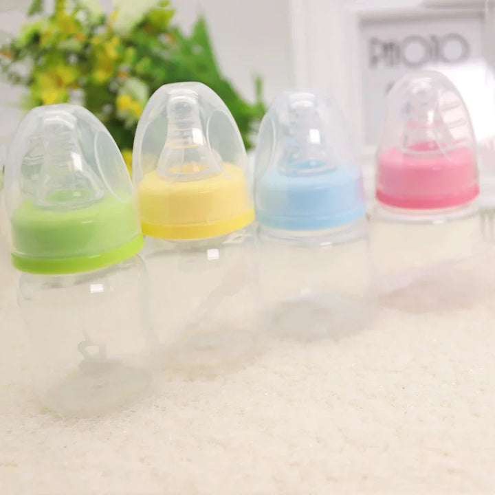 Four clear baby bottles with green, yellow, blue, and pink caps on a soft surface with blurred flowers and photo frame background
