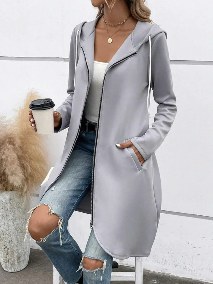 Woman wearing light gray zip-up hooded cardigan with pockets, white tank top, and ripped blue jeans holding coffee cup