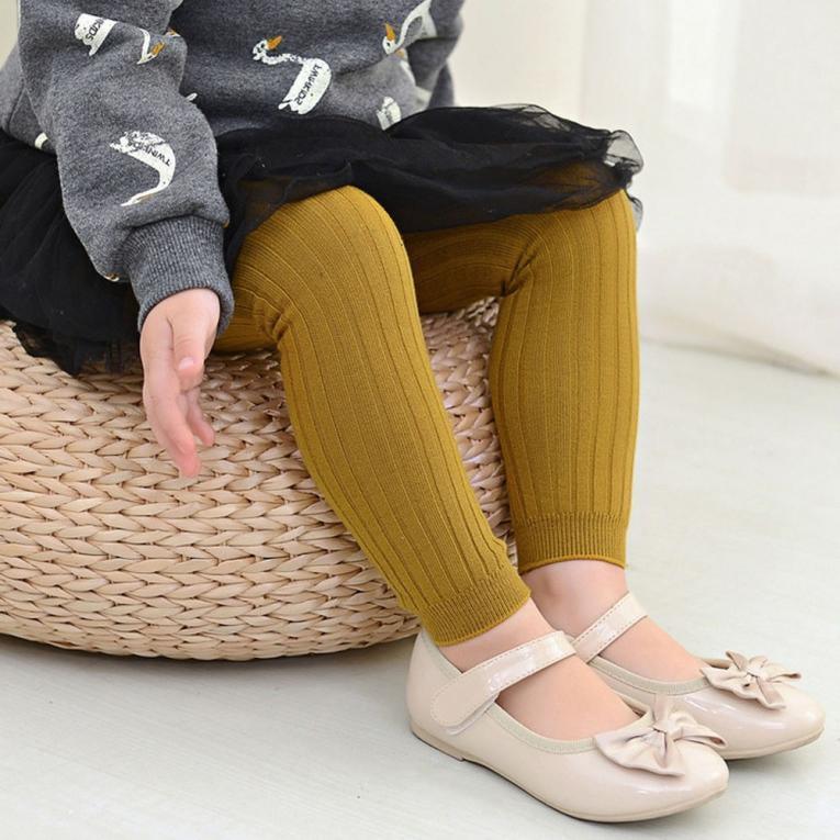 Child wearing mustard knitted leggings, beige bow shoes, and a swan print grey sweater sitting on a woven pouf