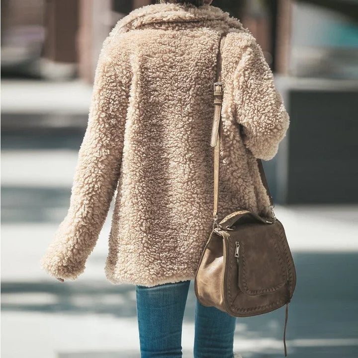 Woman wearing beige teddy coat and blue jeans carrying a brown leather shoulder handbag outdoors
