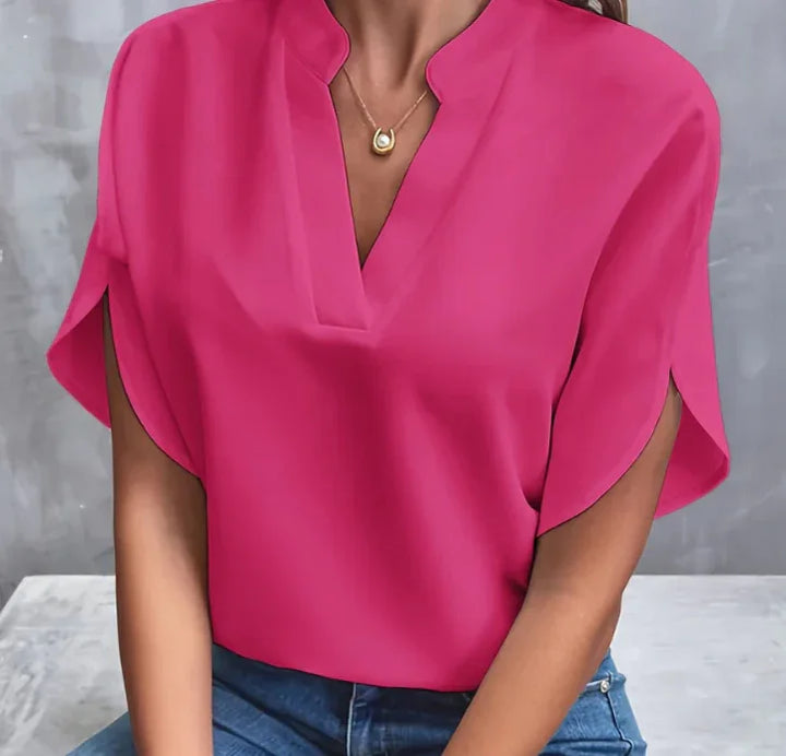 Woman wearing bright pink short-sleeve blouse with V-neck and gold pendant necklace, paired with blue jeans