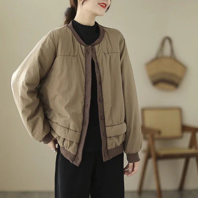 Woman wearing a casual beige button-up padded jacket with brown trim over black outfit in minimal indoor setting
