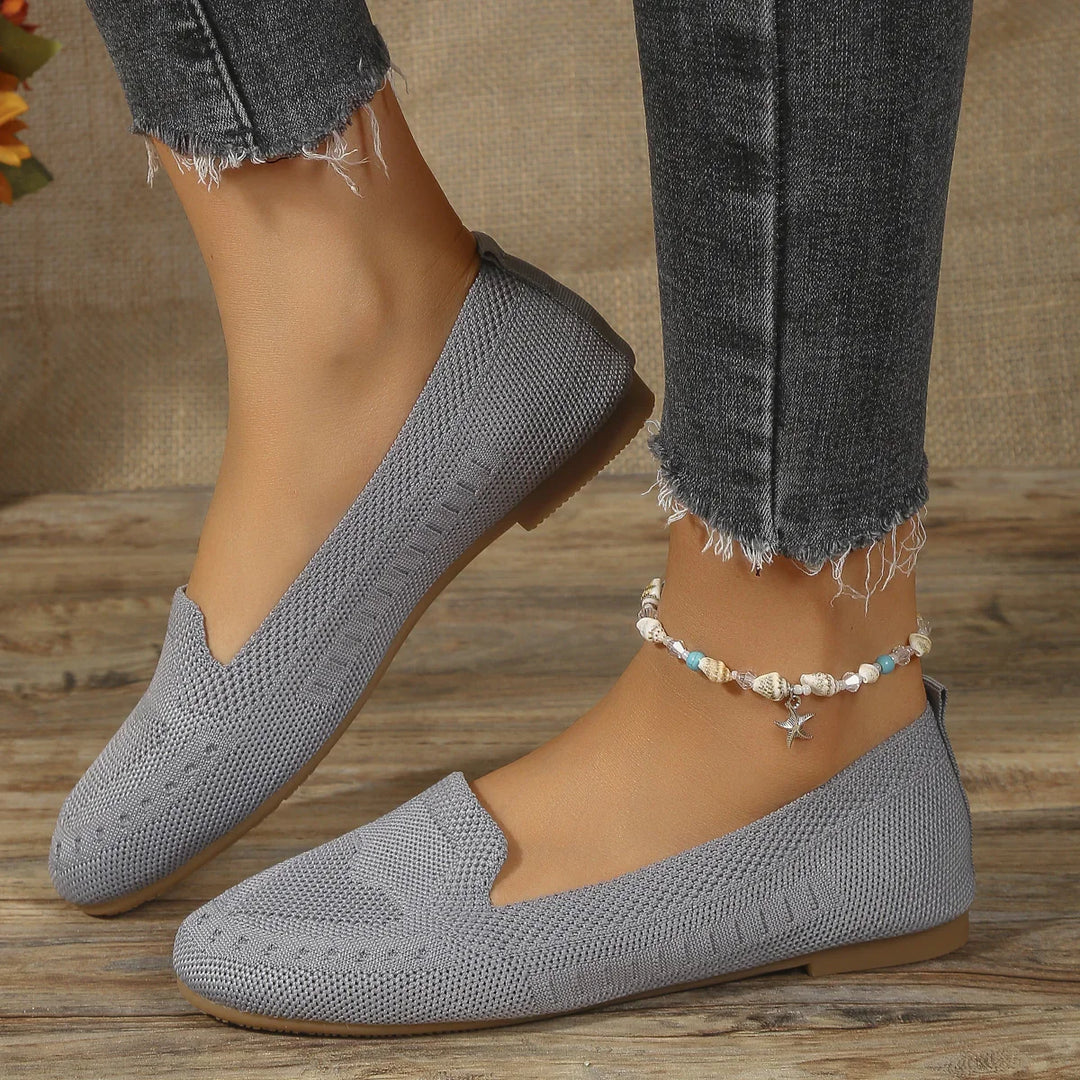 Close-up of feet wearing gray knit slip-on shoes and a seashell anklet, paired with frayed black jeans