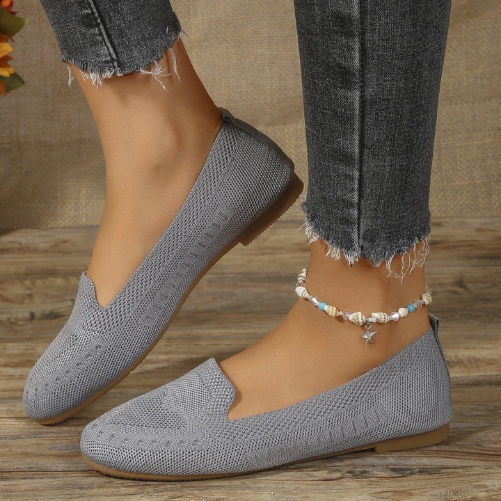 Close-up of feet wearing gray knit slip-on shoes and a seashell anklet, paired with frayed black jeans