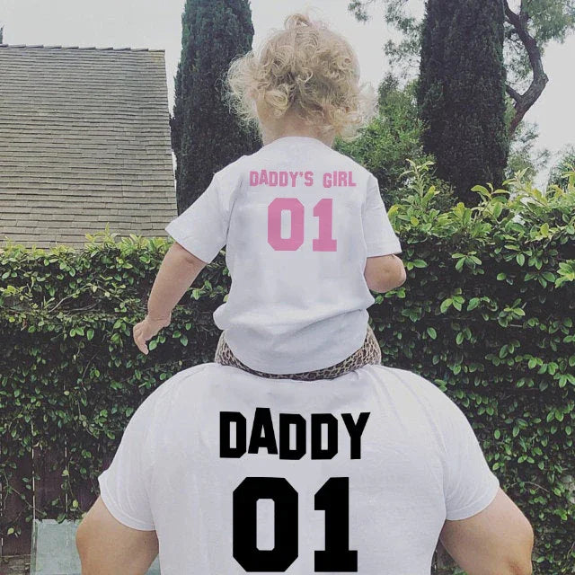 Father carrying toddler girl on shoulders, both wearing matching white shirts with 'Daddy 01' and 'Daddy's Girl 01' printed