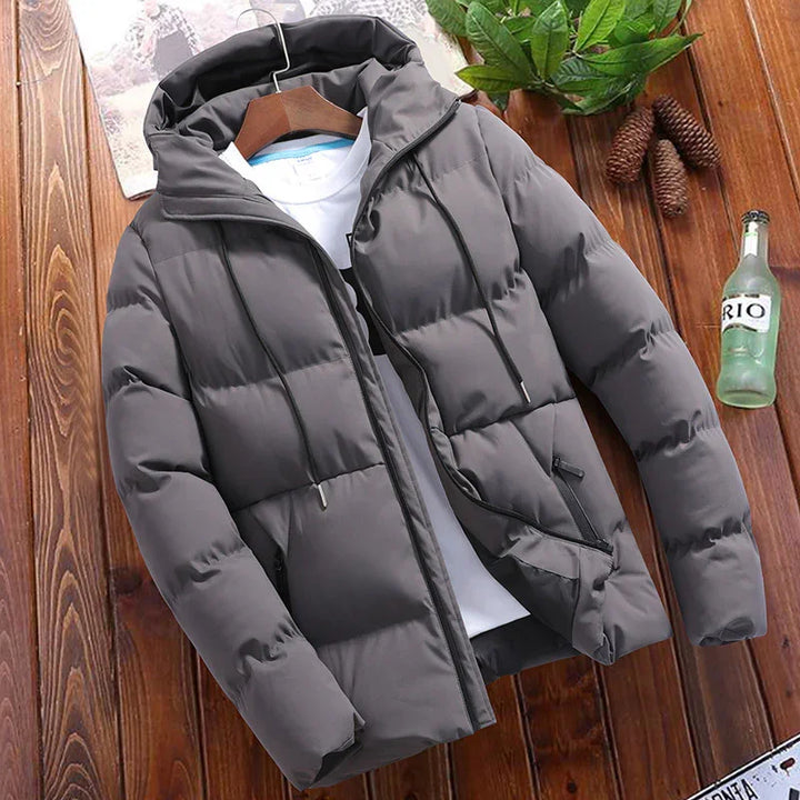 Gray padded hooded winter jacket with zipper and pockets on wooden surface with green plant and glass bottle