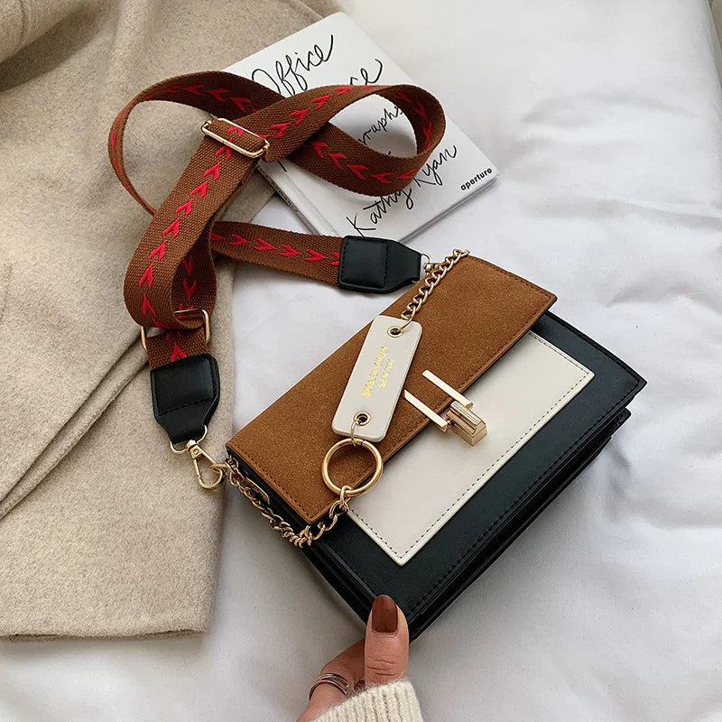 Color block crossbody handbag with brown suede, cream and black panels, gold lock, and red-patterned strap on beige fabric