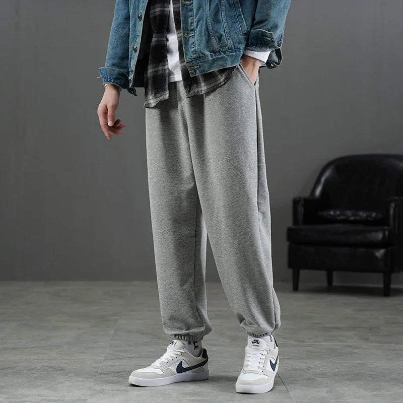 Man wearing grey jogger sweatpants, white Nike sneakers, denim jacket, and checked shirt in indoor setting