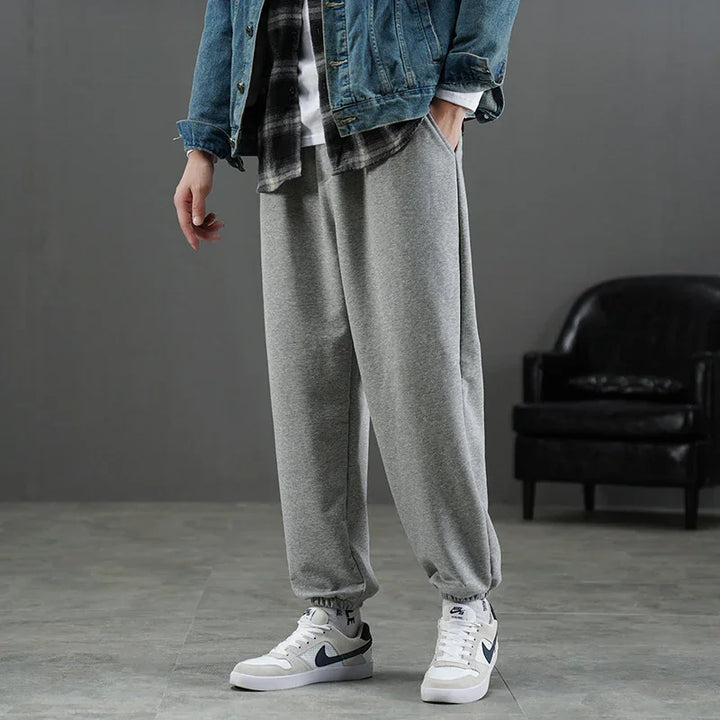 Man wearing grey jogger sweatpants, white Nike sneakers, denim jacket, and checked shirt in indoor setting
