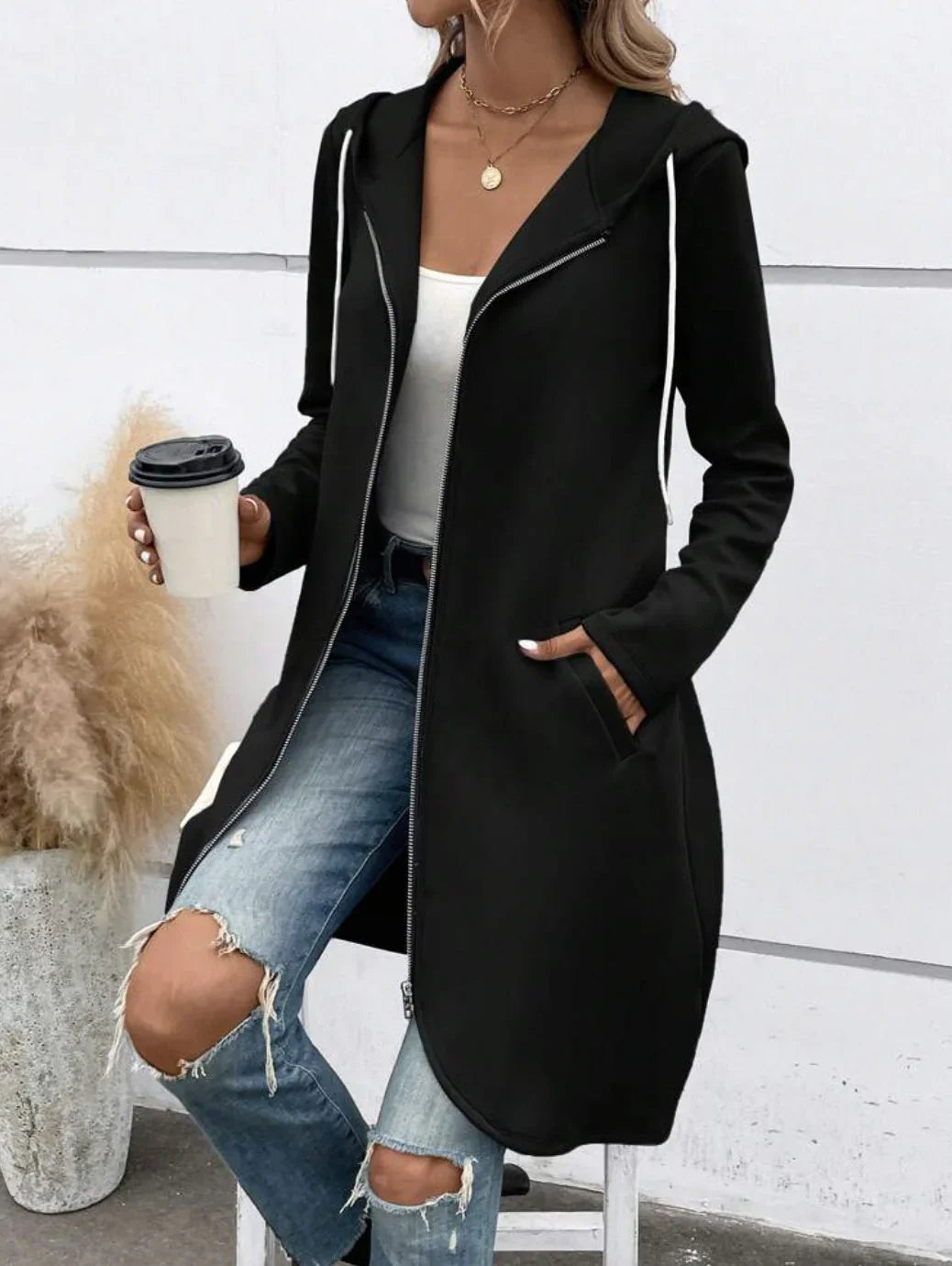 Woman in black zip-up hoodie coat, white tank top, ripped blue jeans, holding takeaway coffee cup