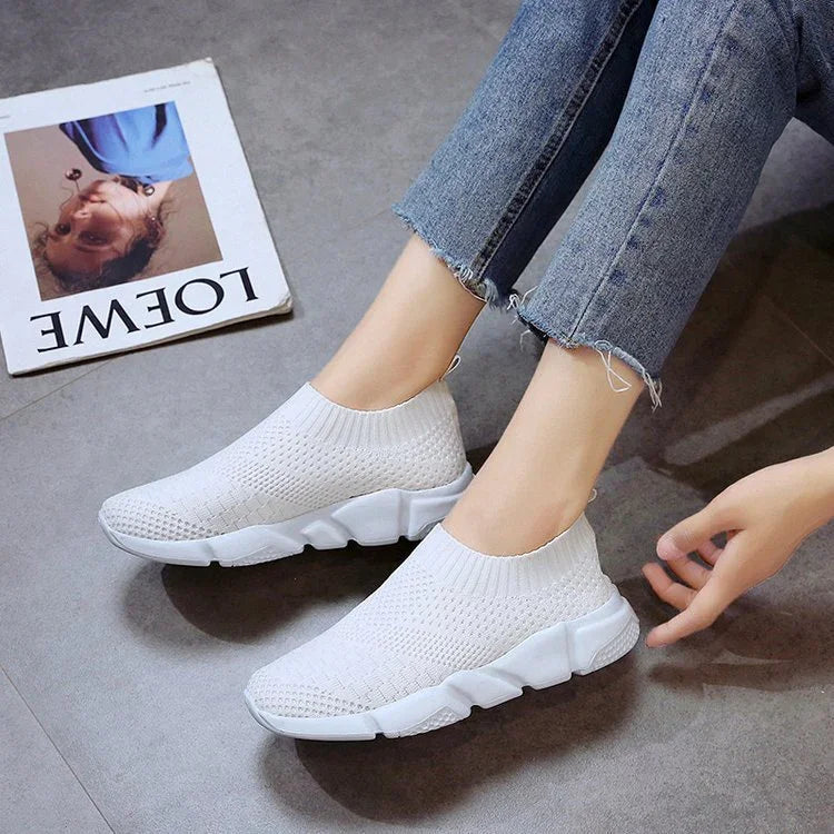 Close-up of white knitted slip-on sneakers on feet with frayed hem blue jeans on gray floor