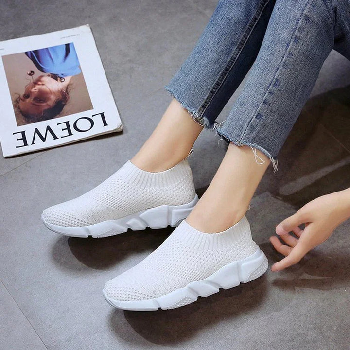 Close-up of white knitted slip-on sneakers on feet with frayed hem blue jeans on gray floor
