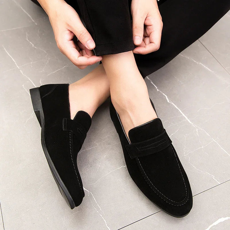 Close-up of person wearing black suede loafers on gray tiled floor, hands adjusting black pants