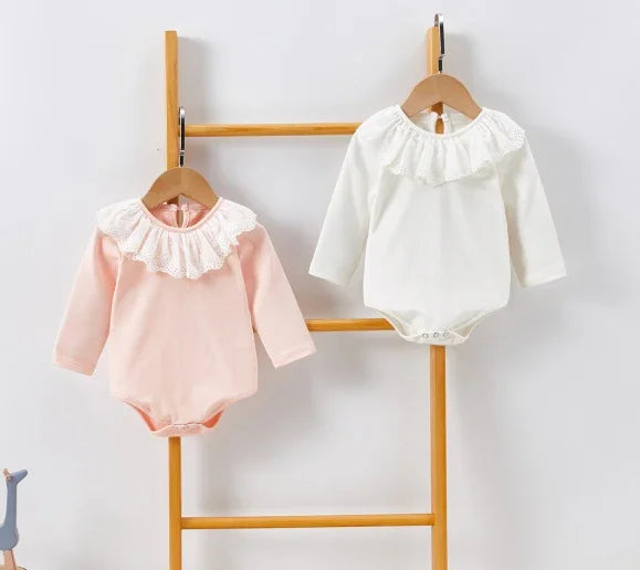 Pink and white baby bodysuits with lace collars hanging on wooden ladder against white wall