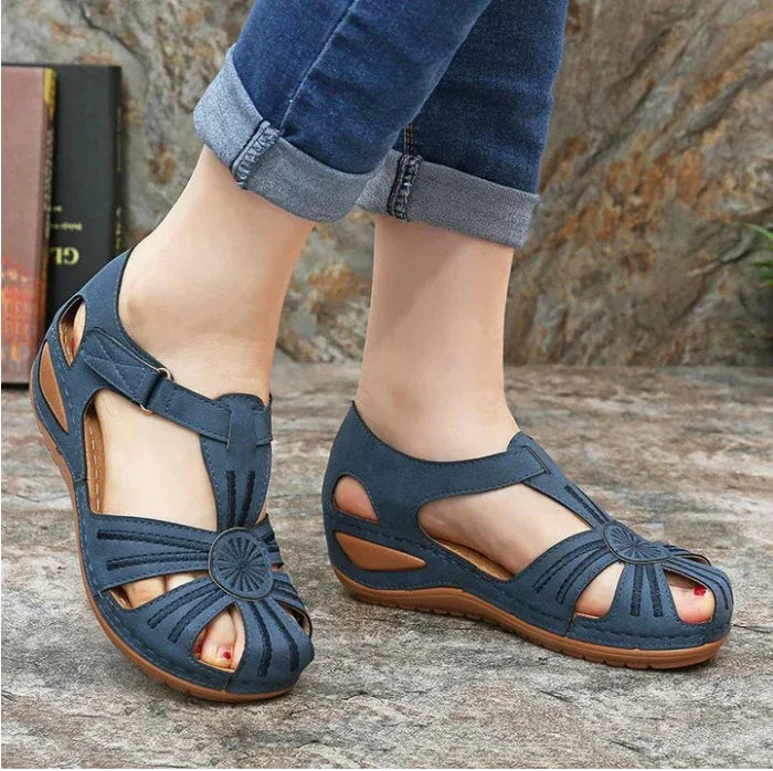 Casual blue women's sandals with decorative cutouts and brown sole worn with cuffed blue jeans on stone floor