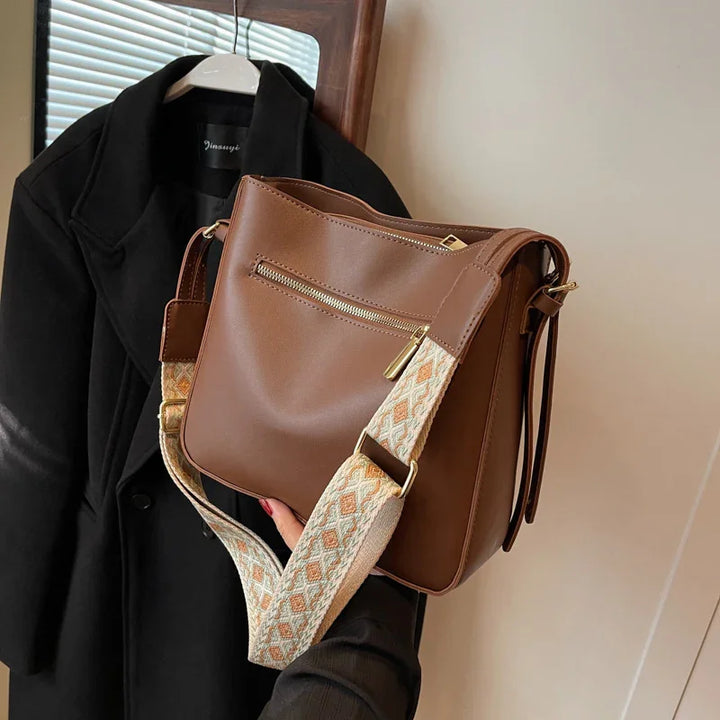 Brown leather handbag with beige patterned strap held in front of hanging black coat and window blinds