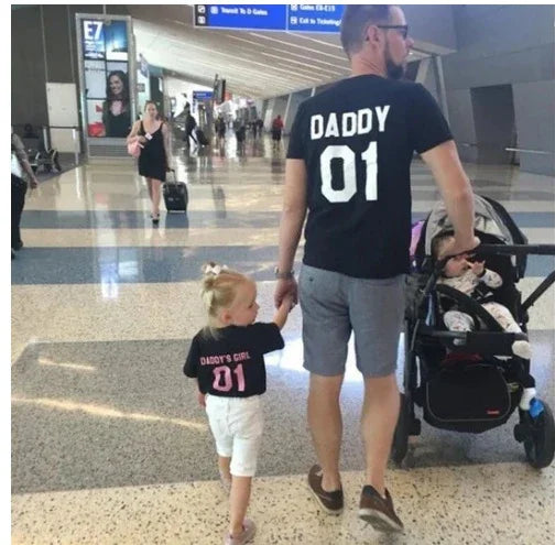 Man wearing black shirt with Daddy 01 and child holding hands wearing Daddy's Girl 01 shirts walking in airport terminal with baby stroller
