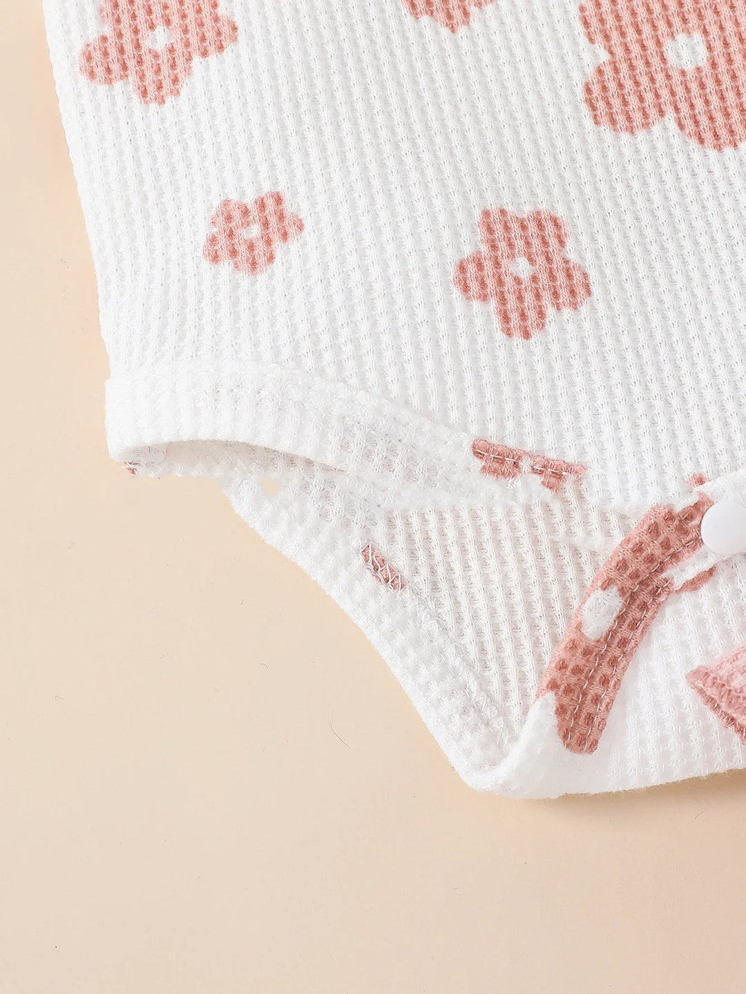 Close-up of textured white baby onesie with pink floral print and snap buttons on beige background