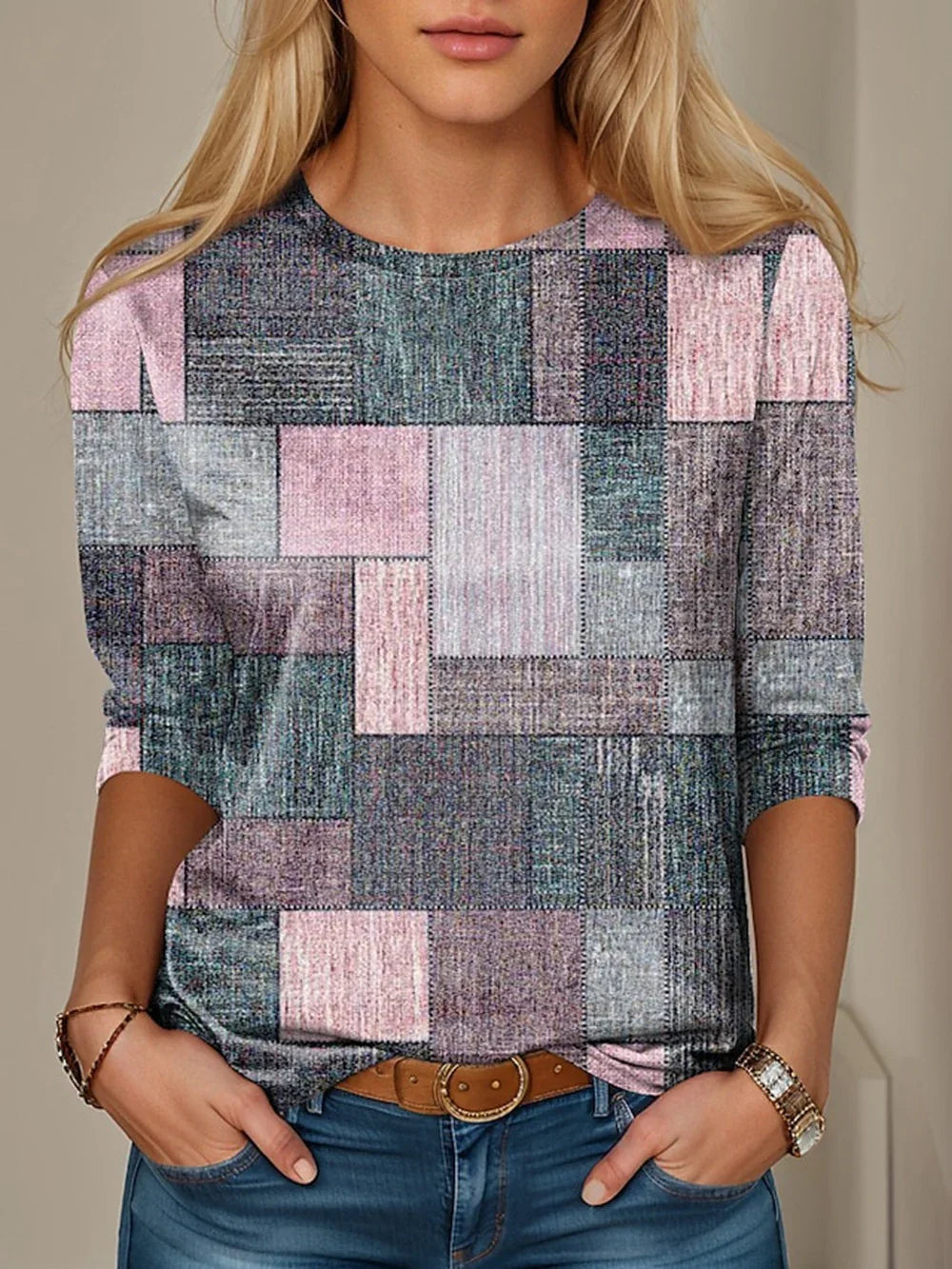 Woman wearing a patchwork pattern long-sleeve top in muted pink, gray, and blue shades with jeans and brown belt