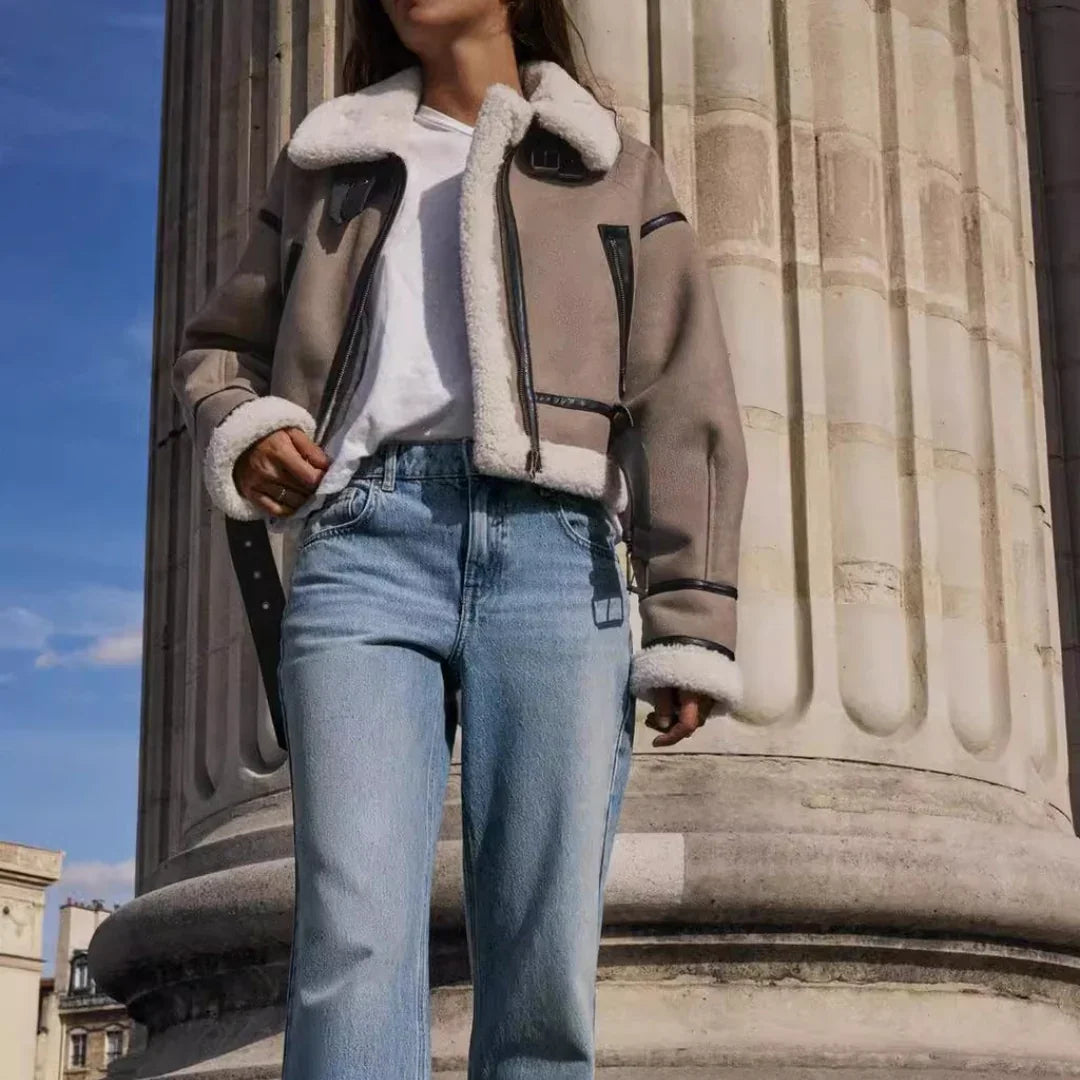 Woman wearing shearling jacket, white t-shirt, and blue jeans posing near stone column under blue sky