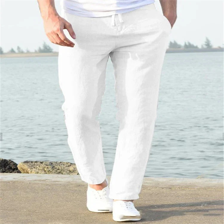 Man wearing casual white linen pants and white sneakers walking near waterfront on sunny day