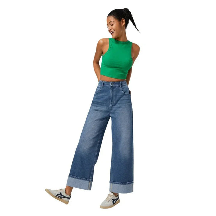 Woman wearing green sleeveless crop top, wide-leg cuffed blue jeans, and white sneakers on white background