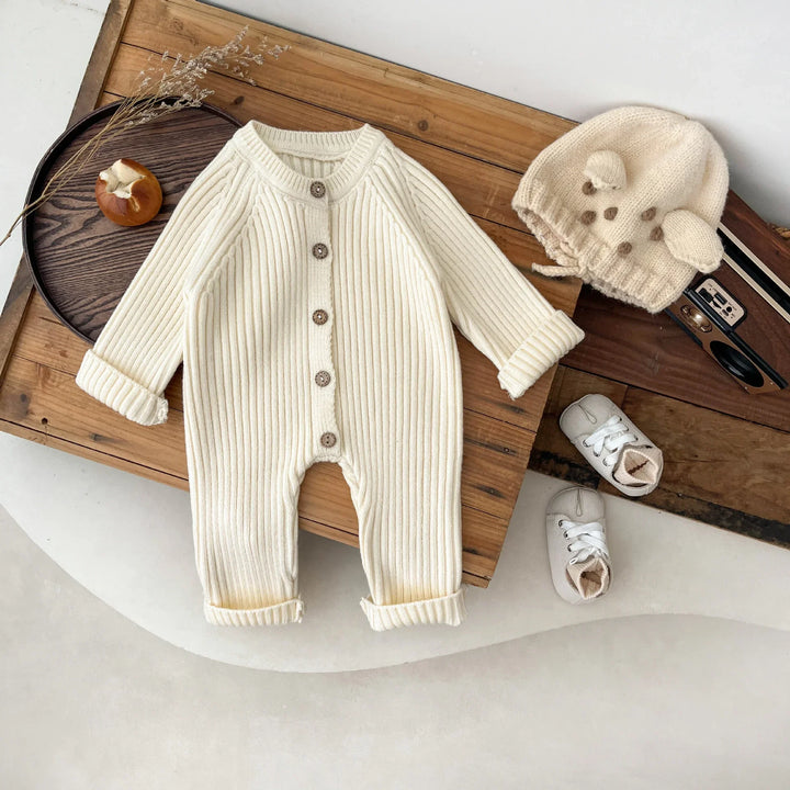 Cream ribbed baby onesie with wooden buttons, knit bear hat, and soft baby shoes on wooden boards