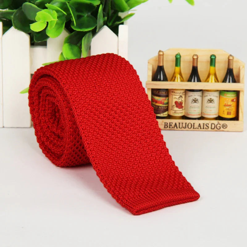 Rolled red knitted tie on white surface with miniature wine bottle crate and green leaves background