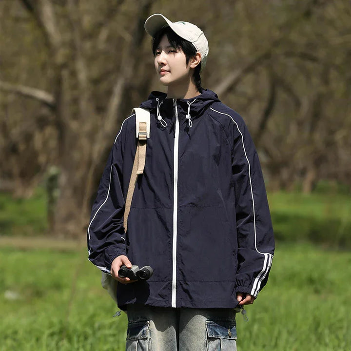 Person wearing navy windbreaker and beige cap holding binoculars in green outdoor setting