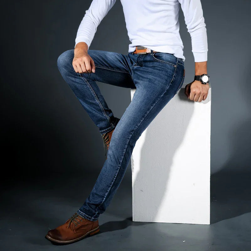 Man wearing slim fit blue jeans, brown leather boots, white long-sleeve shirt, and watch sitting on white block