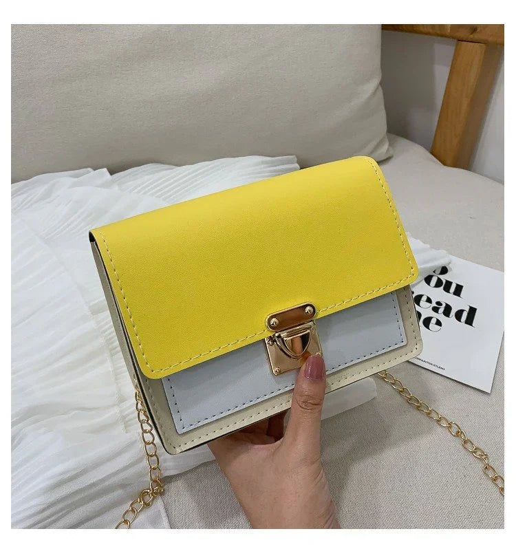 Hand holding small color block crossbody bag with yellow flap, light gray front, gold clasp, and chain strap