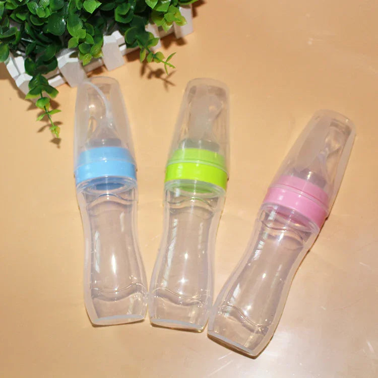 Three baby feeding bottles with colorful caps in blue, green, and pink on a beige surface next to green plant leaves