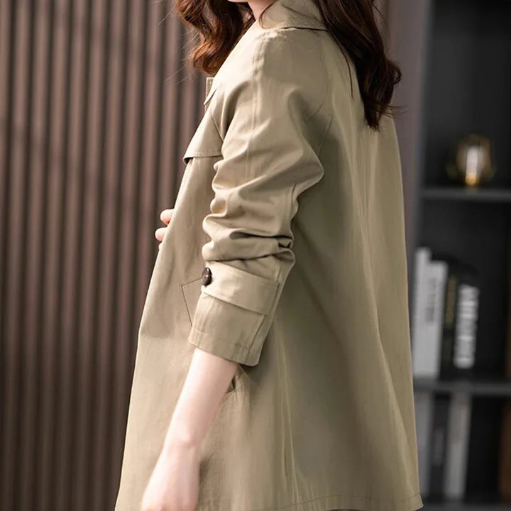 Woman wearing beige trench coat with rolled-up sleeves standing indoors by bookshelf