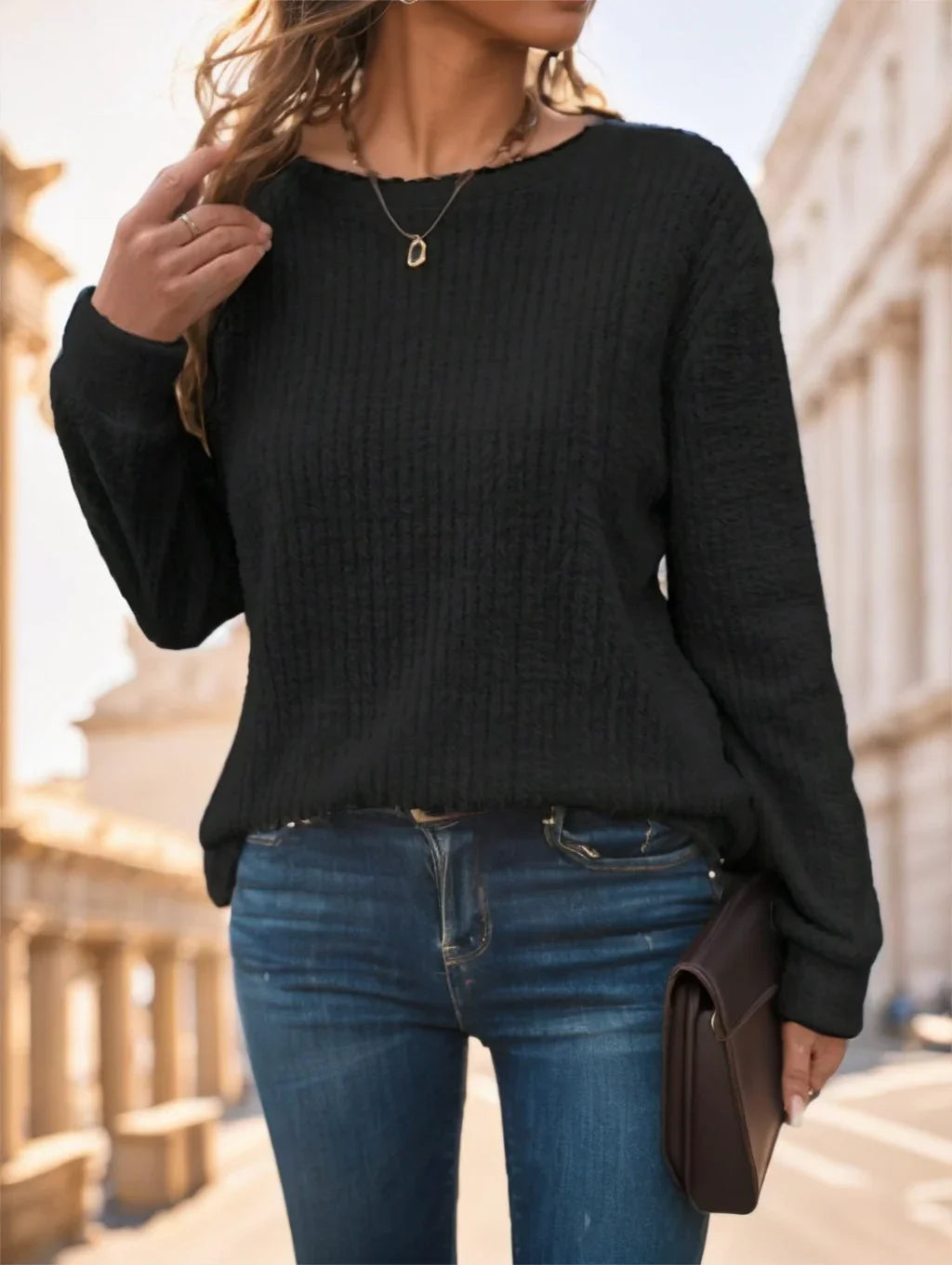 Woman wearing black textured sweater and blue jeans holding brown clutch outdoors