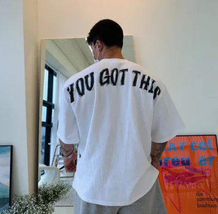 Man wearing white T-shirt with bold text 'YOU GOT THIS' standing in a bright room with wall art and mirror