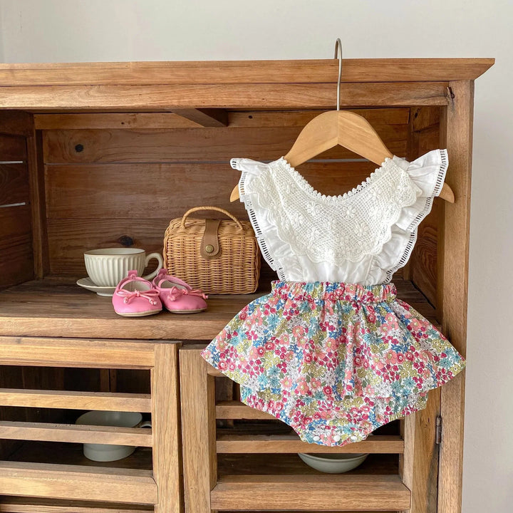 White lace baby romper with floral skirt hanging on wooden cabinet next to pink baby shoes and wicker purse
