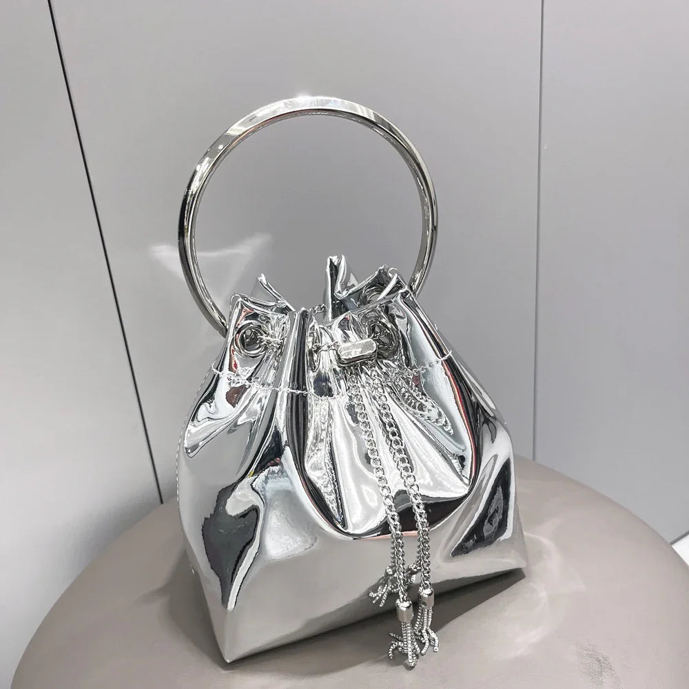 Shiny silver metallic handbag with circular handle and chain drawstring on gray surface
