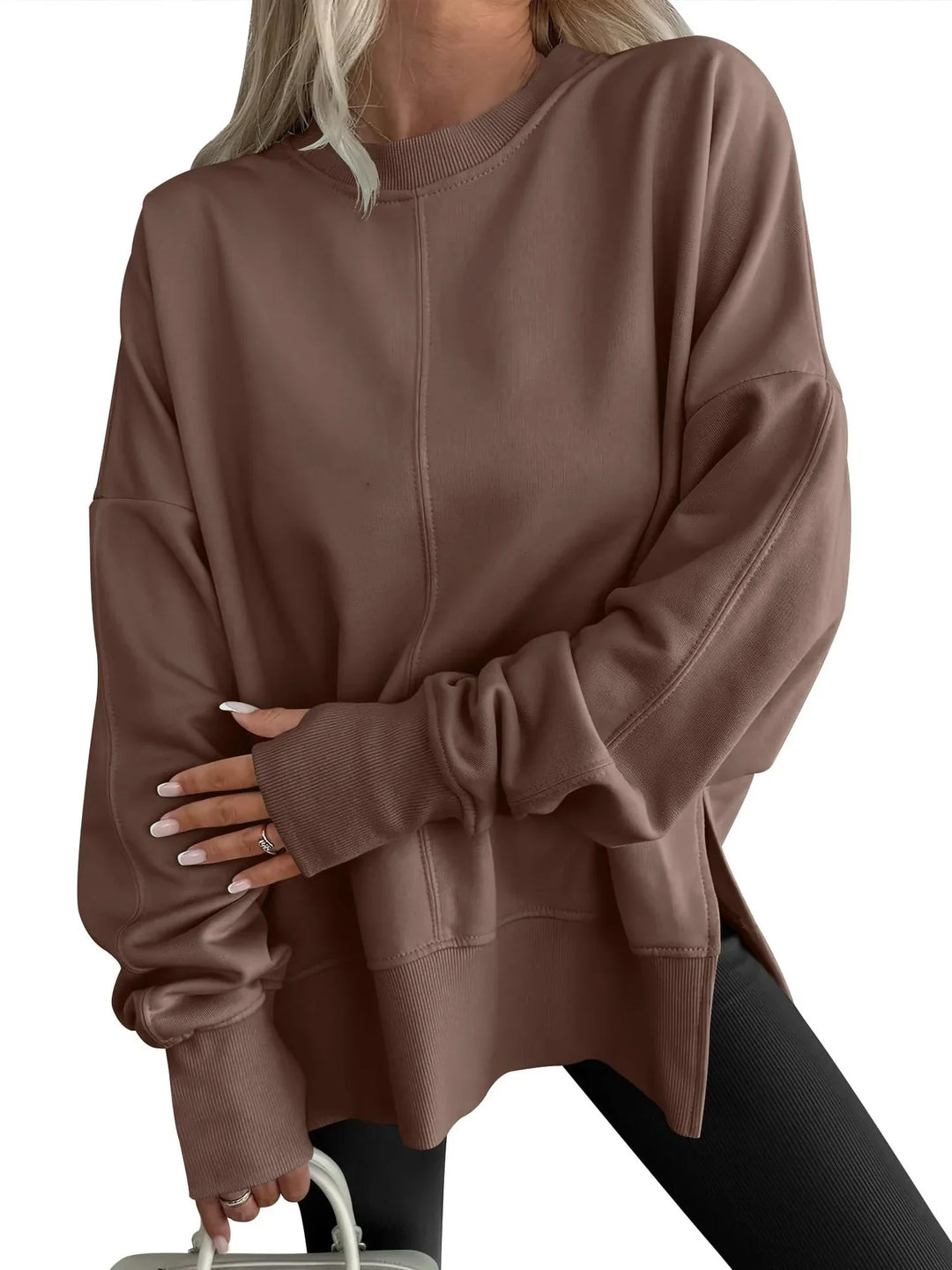 Woman wearing oversized brown sweatshirt with ribbed cuffs and black pants holding white handbag