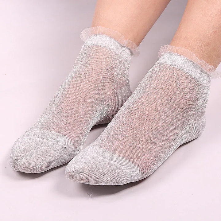 Pair of feet wearing sheer silver glitter socks with ruffled lace cuffs on a light background